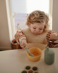 A young child in a striped shirt and bib eats fruit from a yellow bowl using a mushie Silicone Feeding Spoon. A green bottle and cookies are also on the table.