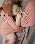 A baby in a mushie Baby Wrap made from organic cotton, wearing a brown knit hat with white spots and ear shapes, is cradled by an adult and holding a green teething toy, looking comfortable and relaxed.