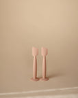 Two pale pink Mushie Diaper Cream Applicators, made of silicone and designed for applying diaper cream, stand upright side by side on a neutral beige surface with a plain beige background.
