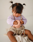 A young child with pigtails, wearing a lavender shirt and beige overalls, looks down while playing with the peach-colored mushie Snack Cup—a spill-proof silicone cup perfect for toddlers.