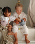 Two smiling children sit on white steps, playing together. One gently reaches into the other’s mushie Snack Cup while they wear cozy clothes and enjoy their time with pastel-colored teapots.
