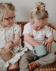 Two young toddlers with light hair sit on a wicker chair. One child, wearing glasses, looks at the other, who reaches into a mushie Snack Cup with handles. Both children are using spill-proof silicone snack cups from mushie.