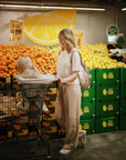 A woman smiles at a small child seated in a shopping cart using a mushie Multi-Use Cover in the grocery store's produce section, surrounded by stacks of lemons and oranges.