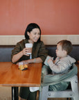 A woman smiles while holding a cup, sitting across from a young child in a high chair at a wooden table, with fruit in a plastic container and the mushie Multi-Use Cover draped nearby, inside a cafe with an orange wall.