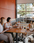 Two women sit at a cafe table with young children; one enjoys a drink, the other sits in a high chair holding a cup. A mushie Multi-Use Cover drapes stylishly over the car seat nearby as sunlight streams through large windows.