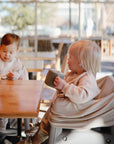 Two young children sit at a wooden table in a sunlit cafe. The mushie Multi-Use Cover drapes over their carseat by the window, adding comfort and function to the cozy scene.
