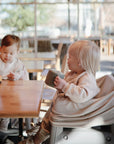 Two toddlers sit at a wooden table in a bright café. One in a high chair drinks from a gray, spill-proof cup, while the other reaches for a mushie Snack Cup. Sunlight pours in through the large windows.