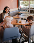 Two women smile over drinks at a cozy café table while two young children in high chairs interact. One child is comfortably covered with the mushie Multi-Use Cover, adding both style and function to the bright, cheerful indoor scene.