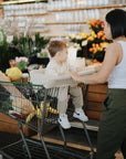 A woman in a white tank top pushes a cart packed with groceries, her toddler seated inside cozy under the mushie Multi-Use Cover, as they pass vibrant flowers in the store.