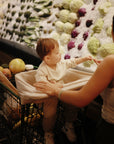 A toddler in a shopping cart reaches for cabbages and other veggies while an adult stands nearby in the grocery store produce section, with a mushie Multi-Use Cover draped over the car seat beside them.