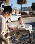 A woman smiles at a baby seated in a shopping cart, cozy in a mushie Multi-Use Cover, on a sunny day. She gently guides the cart while palm trees and cars are visible in the background.