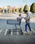 A woman pushes a shopping cart with a child seated inside, cozy under the mushie Multi-Use Cover, across a sunny parking lot with trees, cars, and buildings in the background.