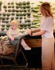A woman, with a young child in light clothes seated in her cart, uses the mushie Multi-Use Cover while shopping in the produce section, with broccoli and cabbage displayed behind them.