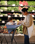 A woman carrying a tote bag smiles at a baby, cozy in a mushie Multi-Use Cover, seated in a shopping cart at the grocery store with fresh vegetables and greens visible on the shelves behind them.