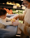 A smiling woman helps a baby in a shopping cart hold a yellow apple in the produce section, with veggies in the background. The baby's carseat is covered with a stylish mushie Multi-Use Cover.