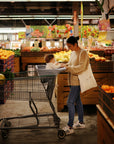 In a grocery store filled with colorful produce, a woman in a beige sweater and blue jeans smiles at a baby seated in the cart, protected by the mushie Multi-Use Cover. She carries a white tote bag.