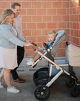 A smiling couple pushes a toddler in a stroller along a brick wall. The child, cozy beneath the mushie Multi-Use Cover, looks at the camera while the adults share a joyful moment together.