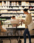 A woman smiles at a baby seated in a shopping cart with the mushie Multi-Use Cover, near a cheese display at the grocery store. She holds a package, with groceries in the cart and a tote bag over her shoulder.