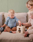 A woman helps two toddlers play with a shape-sorting toy on a beige couch. A mushie Snack Cup sits nearby, its spill-proof silicone design keeping snacks tidy while the children enjoy their activity.