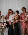 Three women, each holding a baby and a mushie Silicone Baby Rattle Toy made from food-grade silicone, stand together indoors against a white background, smiling and dressed in stylish casual wear.