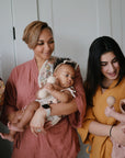 Two women, each holding a baby, smile side by side. One baby uses a pacifier while the other enjoys the mushie Silicone Baby Rattle Toy made from food-grade silicone. All wear warm-toned outfits against a white background.
