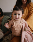 A smiling baby in a pink knitted romper stands holding a blush pink cloth and the mushie Silicone Baby Rattle Toy, creating a fun sensory experience. In the background, two women sit nearby—one in a yellow robe, one holding a baby.