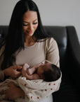 A woman with long dark hair, in a beige top, sits on a dark couch gently cradling her newborn wrapped in the mushie Organic Cotton Muslin Swaddle Blanket, smiling tenderly down at the baby.