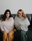 Two women sit on a gray couch, each with a mushie Multi-Use Cover draped around their shoulders. One smiles widely and wears glasses; a baby bottle sits between them.