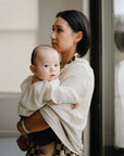 A woman with dark hair holds a baby wearing a white muslin outfit, gazing out the window. The baby is wrapped in the mushie Multi-Use Cover and looks at the camera with their reflection visible in the glass.