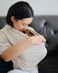 A woman with short dark hair sits on a couch, looking down as she adjusts a light-colored mushie Multi-Use Cover around her torso, which can also be used as a nursing cover.