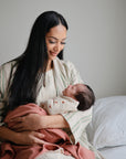 A woman with long dark hair sits on a bed, gently cradling her newborn wrapped in a mushie Organic Cotton Muslin Swaddle Blanket. She smiles softly as she gazes lovingly at her baby.