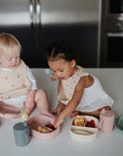 Two toddlers sit on a kitchen counter, wearing bibs and snacking from mushie Lunchboxes—pastel, non-toxic plastic containers with built-in compartments holding finger foods like fruit and pancakes.