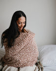 A woman with long dark hair sits on a bed, wrapped in a mushie Multi-Use Cover in brown and white leopard print, smiling gently. The background is simple with white pillows and bedding.