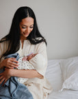 A woman with long dark hair sits on a bed, smiling down at her newborn, who is wrapped in a mushie Organic Cotton Muslin Swaddle Blanket and wearing a hat. The serene scene features white bedding and a neutral background.