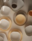 A top-down view of assorted mushie Dinnerware Cups in beige and off-white, non-toxic and in various sizes, scattered on a light surface.