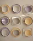 Nine mushie Square Dinnerware Bowls, made from BPA-free plastic in pastel yellow, lavender, and cream, are arranged in a 3x3 grid on a neutral surface with sunlight streaming diagonally across the scene.