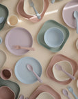 Mushie Dinnerware Cups in soft blue, pink, green, and beige are neatly arranged on a light surface. Some non-toxic cups feature the embossed mushie logo.