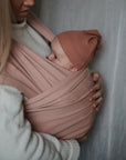 A woman cradles her baby in a mushie Baby Wrap made of soft pink organic cotton, with the baby in a matching hat nestled securely against her chest as she supports the little one with both hands.