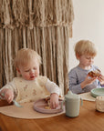 Two young children sit at a wooden table eating snacks. The toddler in front uses a fork with a divided plate on a mushie Silicone Placemat, while the older child eats toast. Soft lighting and textured wall decor complete the cozy scene.