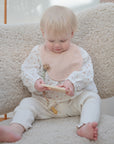 A blond toddler sits on a soft armchair, wearing a pale pink mushie Muslin Bib made from organic cotton and white clothes, looking down while playing with a wooden toy.
