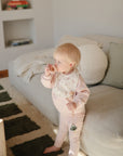A blonde toddler in light pink clothes stands by a beige sofa, wearing a mushie Muslin Bib and holding a pacifier clip, with a pacifier in their mouth, in a softly lit, cozy living room.