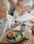 A young blond child eats pastry and banana pieces from a sectioned plate, with mushie Dinnerware Cups nearby. In the background, an adult assists a baby in a high chair.
