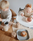 A young boy and a baby sit at a wooden table eating snacks. The baby, wearing a bow headband and bib, eats from a bowl while mushie Silicone Placemats, cups, and plates are neatly arranged on the table.