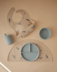 The mushie Silicone Placemat set is displayed on a tan background with a floral-patterned bib, matching food-grade silicone placemat with sections, blue bowl, and sippy cup featuring delicate blue flower designs.