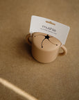 The mushie Snack Cup is a beige, spill-proof silicone cup with two handles and a petal-style opening, ideal for toddler snacks. It sits on a brown textured surface.