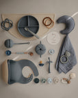 A flat lay of baby and toddler items in soft blue, beige, and gray, featuring a mushie Snack Cup along with a silicone plate, bib, spoons, rattles, teethers, pacifiers, crescent pillow, beaded clips, and a "2 months" milestone card.