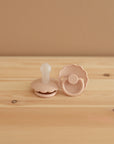 Two light pink FRIGG Daisy Silicone Pacifiers rest on a light wood surface against a beige wall; one stands upright and the other lies flat. This 2-pack from FRIGG is made in Denmark from medical grade silicone with scalloped edges.