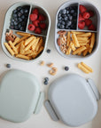 Two mushie Lunchboxes with compartments are filled with blueberries, raspberries, pecans, walnuts, cashews, and tube-shaped snacks. Two light-colored lids are beside them on a white surface. Made from non-toxic plastic for safe snacking.