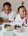 Two smiling children in white shirts relax on a white surface beside mushie Lunchbox containers filled with blueberries, raspberries, and snacks. They look happy enjoying their healthy food from the built-in compartments together.
