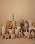 A selection of mushie pastel items—including mugs, baby toys, stacking cups, a Multi-Use Cover, and teething rings—arranged on a beige background in soft pink, green, and cream hues.